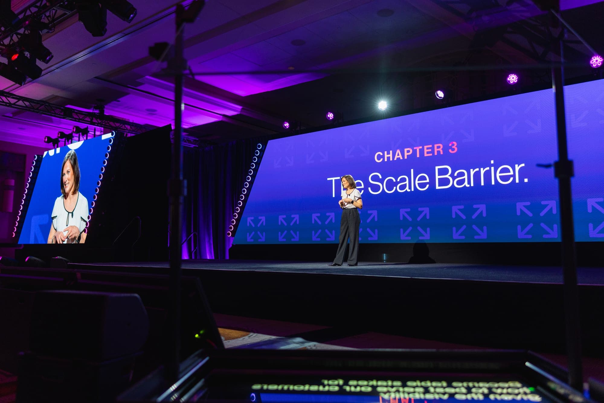 A speaker on the main stage in front of a Chapter 3, The Scale Barrier slide
